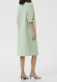 Robe ample vert clair avec manches courtes bouffantes et tissu texturé, se terminant juste au-dessus des chevilles. Associée à des mules nude à talons hauts.