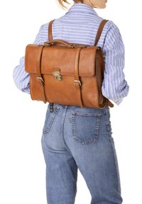 Zaino in pelle marrone con flap frontale, hardware in metallo, due spallacci e dettagli cuciti. Indossato sulla schiena sopra una camicia a righe blu.