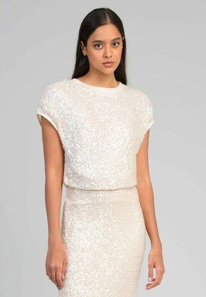 Jeune femme aux longs cheveux noirs portant un haut blanc sans manches orné de sequins et une jupe assortie, sur un fond gris uni.