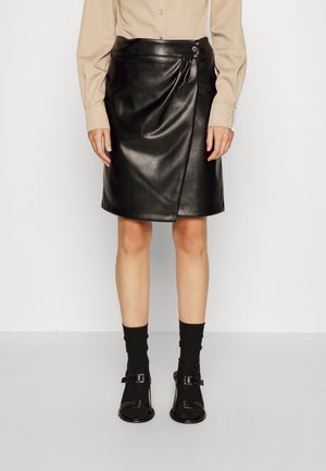 Personne portant une jupe portefeuille en cuir noir avec des détails de boutons, une chemise beige à manches longues, des chaussettes noires et des chaussures noires à boucle, debout contre un mur blanc.