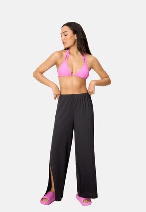 Mujer ajustándose unos pantalones negros de pierna ancha con aberturas laterales, lleva un top de bikini rosa y sandalias de dedo rosas a juego, sobre un fondo liso.