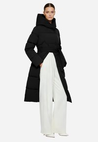 Giacca piumino nera con un grande cappuccio, cintura e sezioni imbottite. Indossata sopra pantaloni bianchi a gamba larga e scarpe bianche a punta.