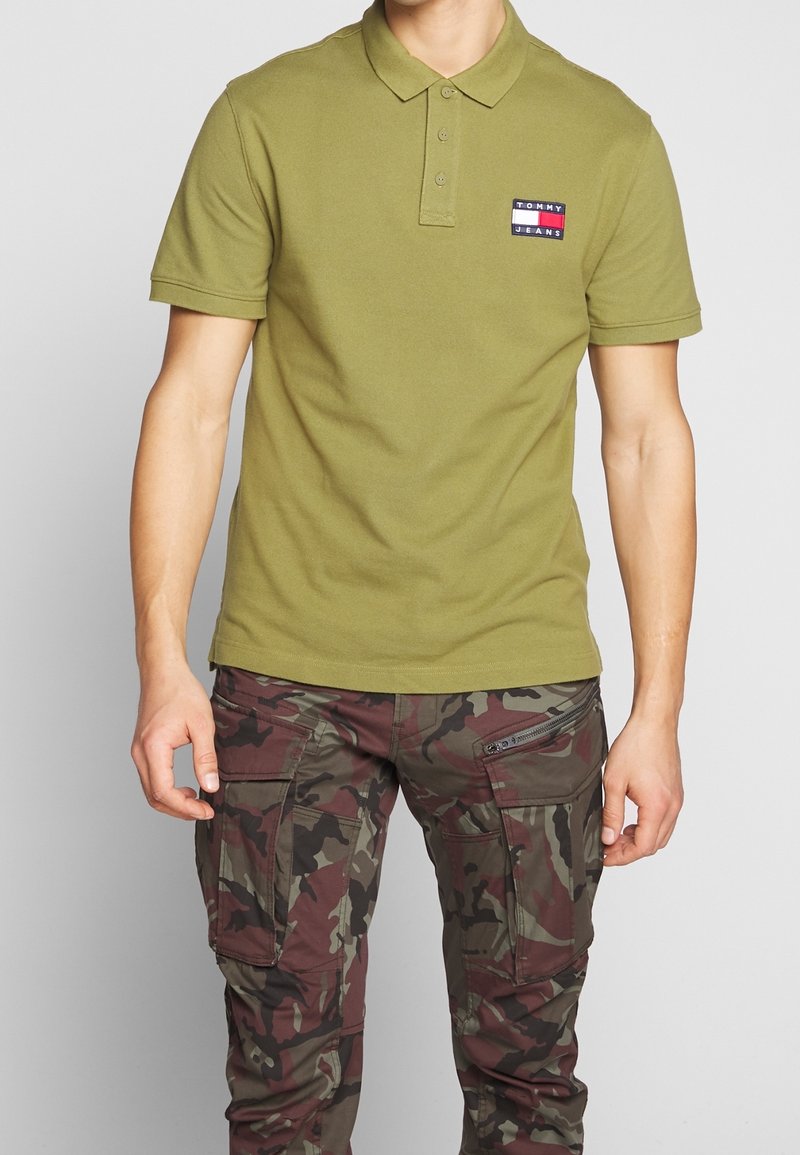 Homme portant un polo Tommy Jeans vert olive et un pantalon cargo camouflage marron-vert sur fond gris uni.