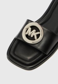 Sort læder slidssandal med en bred rem, der har en cirkulær sølvlogo-accent. Firkantet tå og minimalistisk design med glat tekstur.