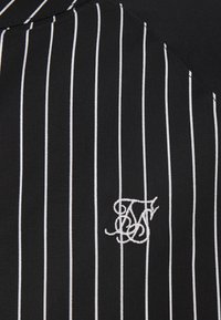 Mustaa kangasta, jossa on pystysuoria valkoisia pinstripeja ja valkoisella brodeerattu koristeellinen monogrammi, joka sisältää kirjaimet T, B ja F.
