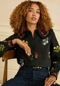 Fekete gombos blúz virágos hímzéssel piros, sárga, kék és rózsaszín színekben. Jellemzője a fodros gallér és a hosszú ujjú, elasztikus mandzsettás ujjak.