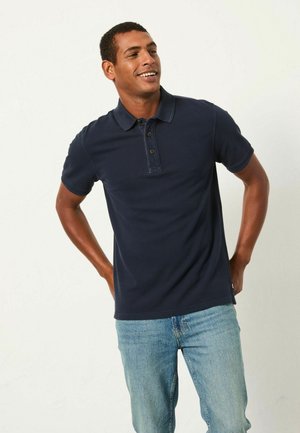 Dunkelblaues Poloshirt aus Baumwolle mit einer Knopfleiste mit zwei Knöpfen und kurzen Ärmeln, kombiniert mit hellblauen Jeans. Einfaches, klassisches Design.