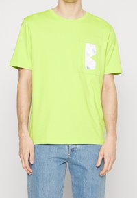 Homme portant un T-shirt vert citron à manches courtes avec un détail réfléchissant argenté brillant sur la poche gauche de la poitrine, associé à un jean bleu clair.