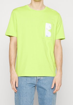 Homme portant un T-shirt vert citron à manches courtes avec un détail réfléchissant argenté brillant sur la poche gauche de la poitrine, associé à un jean bleu clair.