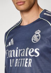 Maglia atletica blu navy con tessuto testurizzato, dotata di accenti verde chiaro, loghi argentati e branding "Emirates LY BETTER."