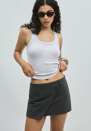 Femme aux cheveux bouclés portant des lunettes de soleil noires, un débardeur gris, des bracelets argentés et une mini-jupe sombre à fines rayures.