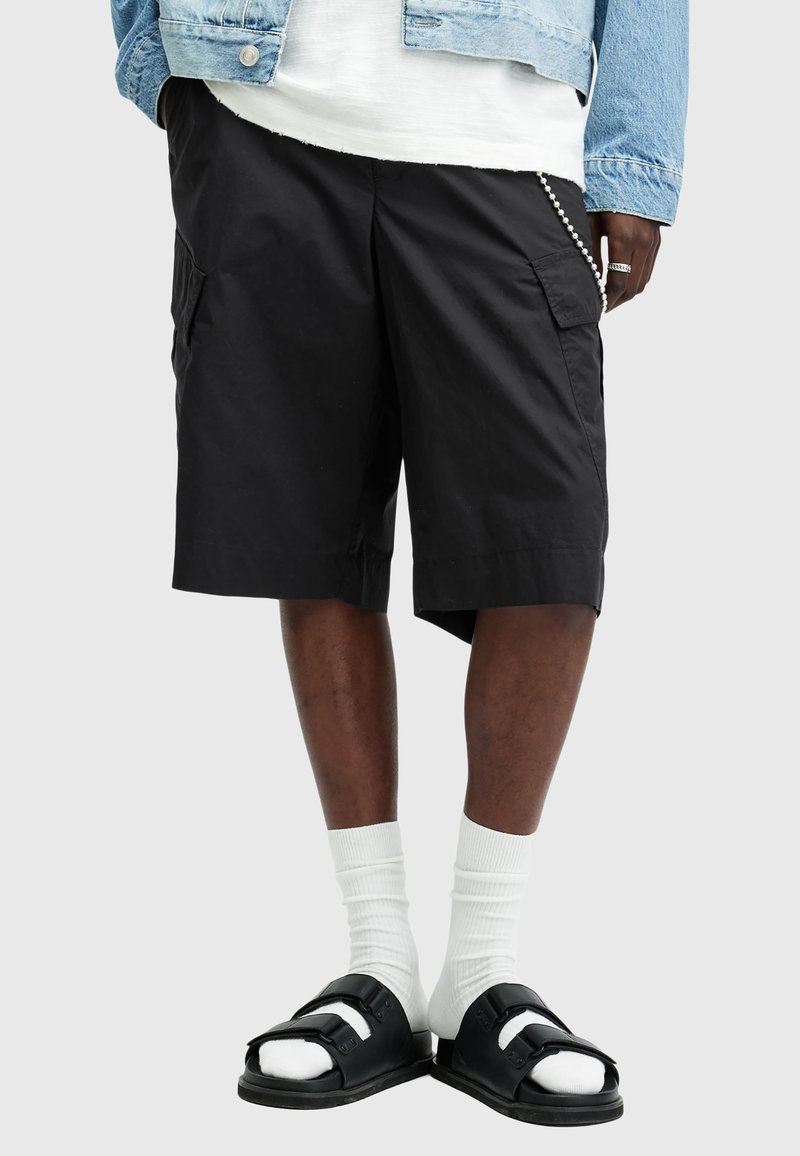 Shorts cargo noirs avec poches latérales, une coupe décontractée et un ourlet jusqu'aux genoux. Portés avec des sandales noires et des chaussettes blanches. Veste en denim superposée.