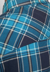 Gros plan sur un tissu à carreaux bleu et marine avec des lignes blanches et grises qui se croisent, montrant des coutures détaillées et la texture.