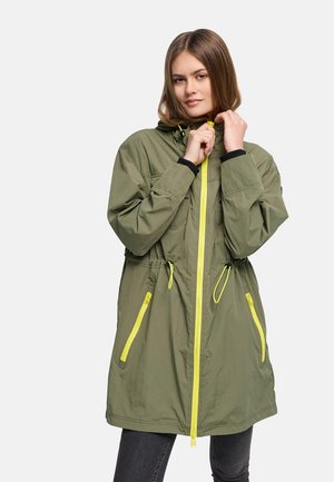 Mlada ženska, ki si prilagaja kapuco parke olivno zelene barve s svetlo rumenim zadrgo in poudarki na žepih, oblečena v temne hlače na enobarvnem ozadju.