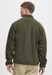 Giacca in pile verde oliva con colletto alto, maniche lunghe e una consistenza morbida. Presenta un orlo arrotondato e un design pulito e semplice.