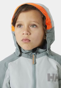 Světle šedá nepromokavá bunda s oranžovou podšívkou kapuce, předním zipem a šedým logem. Má vysoký límec a viditelné švy.