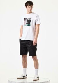 Hvit t-skjorte med et svart og fargerikt grafisk trykk, paired med svarte shorts, hvite sokker og sneakers med svarte detaljer.