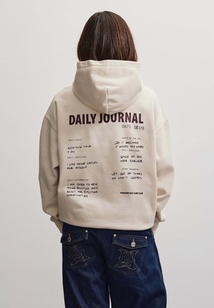 Persona con capelli castani lunghi che indossa una felpa con cappuccio beige con il testo "Daily Journal" e note scritte a mano sulla schiena, abbinata a jeans blu scuro.