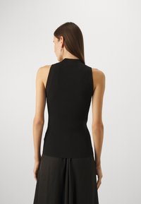 Femme aux longs cheveux bruns portant un top noir côtelé sans manches et une jupe noire, vue de dos sur un fond clair uni.