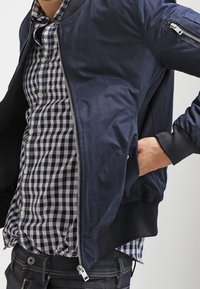 Giacca bomber navy con una texture liscia, polsini a coste e una tasca con zip sulla manica sinistra, indossata sopra una camicia a scacchi bianca e nera.