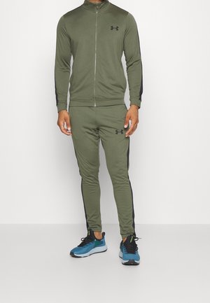 Homme portant un survêtement Under Armour vert olive avec des bandes latérales noires et des chaussures de course bleues, debout devant un fond blanc.