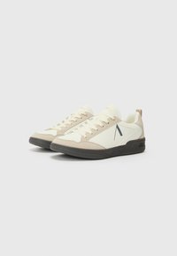 Et par beige og hvite lave sneakers med svarte såler og en svart skrå logo på siden, satt mot en enkel hvit bakgrunn.