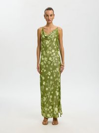 Frau steht und trägt ein ärmelloses, olivgrünes Maxikleid mit floralem Muster, dünnen Trägern und beigen Sandalen vor einem schlichten weißen Hintergrund.