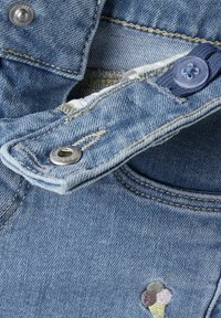 Tela de denim en azul claro con costuras amarillas en contraste. Presenta un botón de presión metálico y un detalle bordado de un cono de helado.