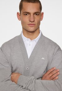 Grå stickad cardigan med V-ringning, med framknäppning och en liten logotyp på vänster bröst, bärs över en vit skjorta med krage.