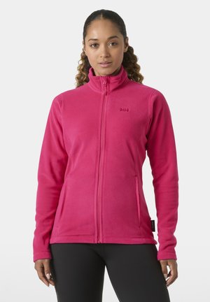 Woman wearing a bright pink zip-up fleece jacket with high collar and black pants, standing against a plain light background.