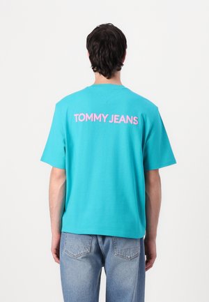 Turkusowa koszulka z krótkim rękawem i okrągłym dekoltem. Na plecach różowy nadruk "TOMMY JEANS". Miękki bawełniany materiał. Swobodny krój.