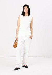 Top blanc sans manches associé à un pantalon large blanc. Le modèle tient un petit sac à main marron et porte des ballerines noires à talons pointus avec des accents décoratifs.