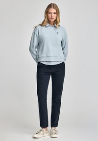 Ljusblå tröja med ribbad mudd, lager över en skjorta med krage, kombinerad med mörka byxor och beige sneakers, stående mot en ljus bakgrund.