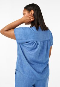 Blå chambray-topp med korta ärmar, med en plisserad bakdesign och en mjuk textur. Stylad med matchande blå byxor.