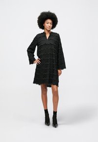 Mulher com cabelo afro, vestindo um vestido preto de manga longa texturizado e sapatos de salto pretos, a posar com uma mão na anca.