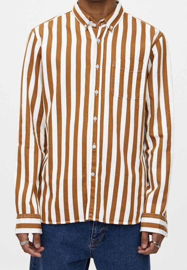 Homme portant une chemise à rayures verticales marron et blanc avec une poche poitrine et un jean bleu, debout les bras détendus.