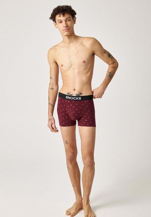 Jeune homme tatoué portant un boxer bordeaux avec une taille noire marquée "SNOCKS", debout pieds nus devant un fond uni.