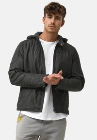 Schwarze Lederjacke mit Kapuze, gesteppte Schulterakzente, Reißverschluss-Taschen und glatter Textur. Getragen über einem weißen T-Shirt und grauen Hosen.