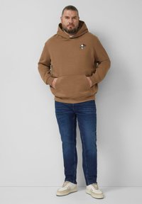 Brązowa bluza z kapturem z przednią kieszenią, ozdobiona małym haftem Snoopy. Do tego niebieskie jeansy i kremowe trampki.