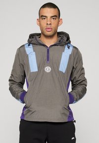 Grå tröja med luva, med blå accenter, dragkedja framtill och en logotyp på bröstet. Tyget verkar lätt och mjukt.