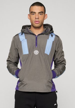 Giacca grigia con cappuccio, con dettagli blu, zip frontale e patch con logo sul petto. Il tessuto sembra leggero e liscio.