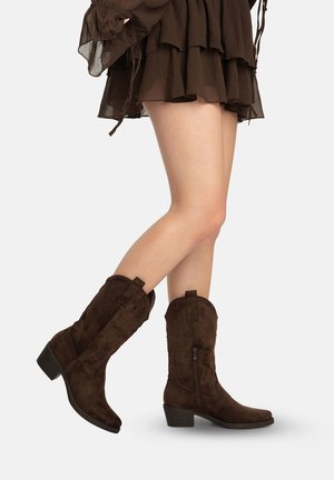 Ben iført mellemstore, brune ruskinds cowboystøvler med dekorative syninger og lave hæle, kombineret med en brun nederdel med flæser.