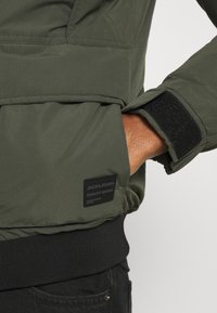 Giacca imbottita verde con etichetta del logo nera, chiusura al polso in velcro e vita elastica. Il tessuto appare liscio e leggero.