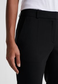 Zwarte op maat gemaakte broek met een gladde textuur, voorzien van zijzakken en een slank ontwerp. Hand rustend op de stof.