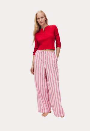 Kvinde stående barfodet iført en rød langærmet cropped top og løse lyserøde bukser med lodrette røde striber.