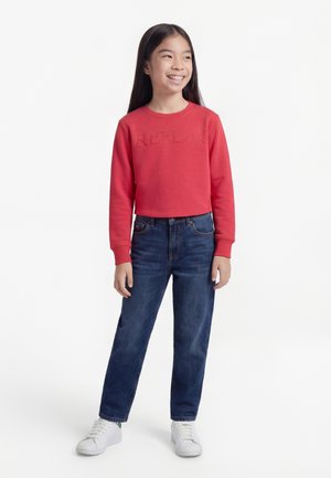 Χαμογελαστή κοπέλα με μακριά μαύρα μαλλιά, φορώντας κόκκινο φούτερ, σκούρο μπλε τζιν και λευκά αθλητικά παπούτσια, στέκεται μπροστά σε λευκό φόντο.