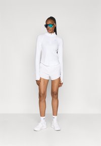 Balta sportinė palaidinė su pusiau užtrauktuku ir ilgomis rankovėmis, derinama su baltais šortais. Modelis dėvi mėlynus akinius nuo saulės ir baltus sportinius batelius.