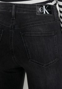 Jeans de mezclilla negra con un ajuste ceñido, diseño de cinco bolsillos y etiqueta con el logo de Calvin Klein en la cinturilla. Detalles de textura sutil.