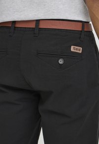 Gros plan d'une personne portant un pantalon noir avec des poches arrière boutonnées et une ceinture marron, avec l'étiquette de marque "Solid" visible sur la poche.