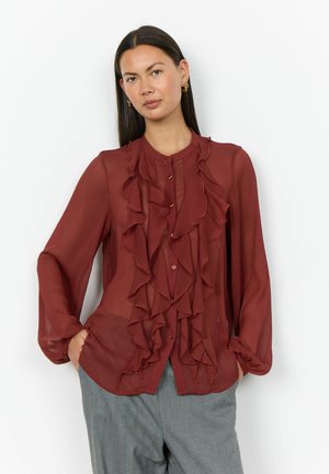 Femme aux longs cheveux foncés porte un chemisier transparent couleur rouille avec des volants et un pantalon gris, debout devant un fond blanc uni.